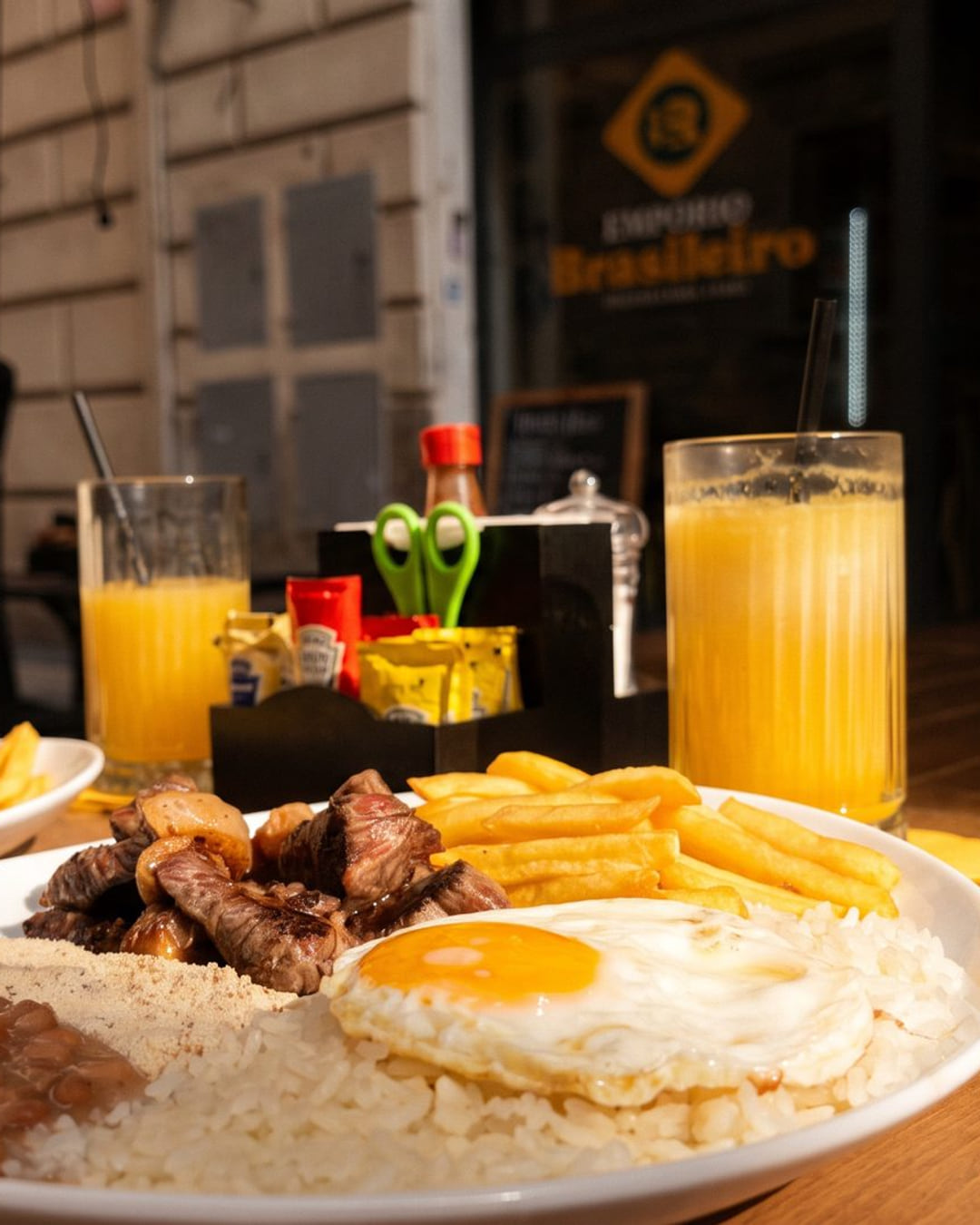 Il prato feito sul tavolo di legno davanti al locale: riso, fagioli, farofa, carne, patatine e uovo, con il succo d'arancia e l'insegna gialla dietro